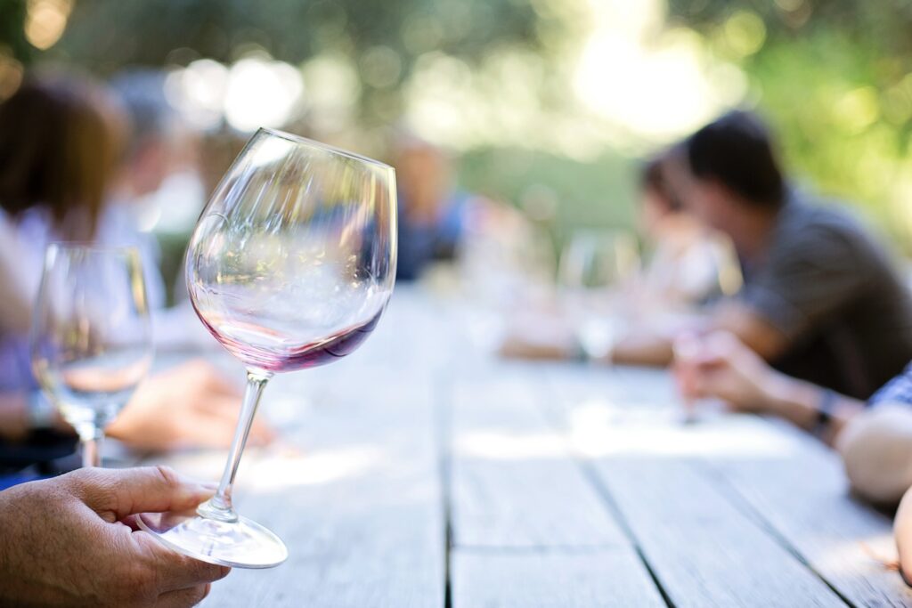 A hand holding a red wine glass at an outdoor dinner party with blurred guests in the background